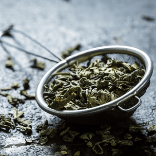 Dried green herbs in a metal strainer on a dark surface